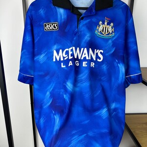 May include: A blue and white football shirt with a black collar and white trim. The shirt has a faded brushstroke pattern and features the Asics logo, the Newcastle United crest, and the text "McEwans Lager".
