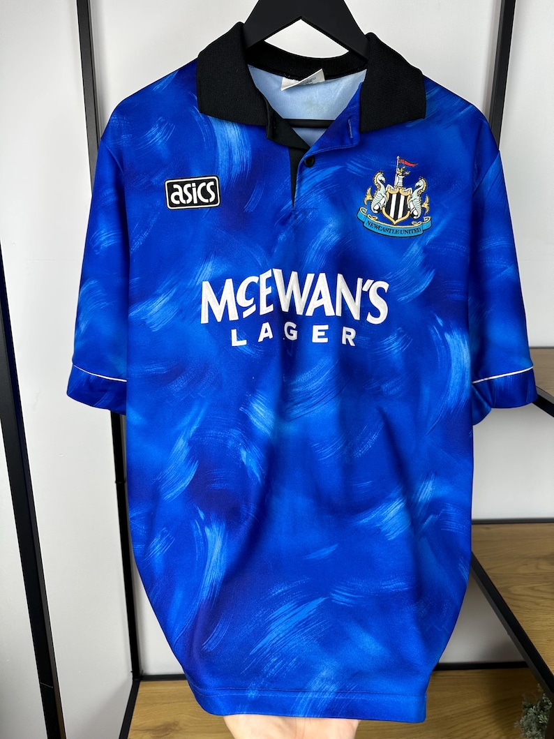 May include: A blue and white football shirt with a black collar and white trim. The shirt has a faded brushstroke pattern and features the Asics logo, the Newcastle United crest, and the text "McEwans Lager".