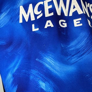 May include: A blue and white football shirt with the text "McEwans Lager" printed on the front.
