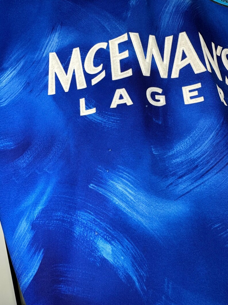 May include: A blue and white football shirt with the text "McEwans Lager" printed on the front.