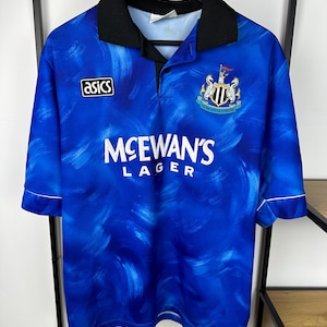 May include: A blue and white football shirt with a black collar and white trim. The shirt features a sponsor logo for McEwan's Lager and the team crest for Newcastle United. The shirt is made by Asics.