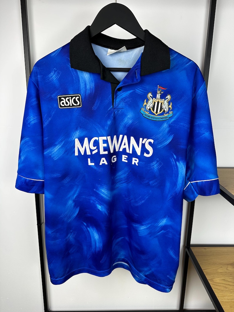 May include: A blue and white football shirt with a black collar and white trim. The shirt features a sponsor logo for McEwan's Lager and the team crest for Newcastle United. The shirt is made by Asics.