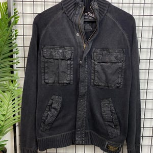 Op de afbeelding: Zwarte gebreide ritsjas met grote cargozakken en een geribbelde kraag. De jas heeft een label met de tekst "Jeans Company" en "L.A. Limited Company".