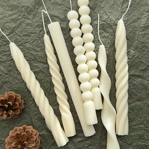 May include: A set of six white candles with different designs, including twisted, beaded, and ribbed. The candles are arranged on a textured green surface.