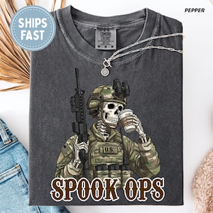 May include: Dark grey t-shirt with a skeleton soldier in camouflage, holding a rifle and a coffee cup. The text "SPOOK OPS" is in a brown font. A silver necklace is draped on the shirt.