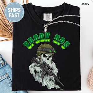 May include: Black t-shirt with a "SPOOK OPS" graphic in green. The design features a ghost wearing a military helmet and holding a rifle. A silver necklace is draped across the shirt.