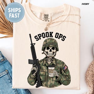 May include: Ivory-coloured t-shirt featuring the text "SPOOK OPS" above a skeleton soldier graphic. The skeleton is wearing a camouflage helmet and uniform, holding a rifle and a coffee cup. The shirt has a crew neck.
