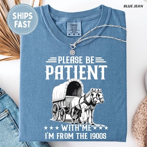 May include: Blue Comfort Colors t-shirt with the text "PLEASE BE PATIENT WITH ME I'M FROM THE 1900S" and a graphic of a covered wagon pulled by two horses. A silver necklace is draped across the shirt.