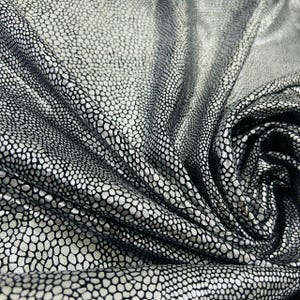 Puede incluir: Primer plano de una tela con un patrón plateado y negro. La tela tiene un diseño texturizado en forma de escamas, con puntos plateados sobre un fondo negro. El material está drapeado, mostrando pliegues y sombras.