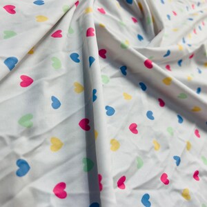 May include: White fabric with a repeating pattern of small, colorful hearts. The hearts are in shades of pink, blue, yellow, and green.