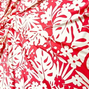 May include: A vibrant red fabric featuring a repeating pattern of large, white tropical monstera leaves and small five-petaled flowers. The material is draped in soft folds, creating a textured surface with a bold, high-contrast botanical design.