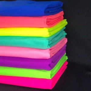 Può includere: Una pila di tessuto dai colori vivaci nelle tonalità di blu, rosa, giallo, verde e viola. Il tessuto è ben piegato, mostrando uno spettro di tonalità vibranti. La disposizione suggerisce un prodotto tessile, forse per l'artigianato o la moda.