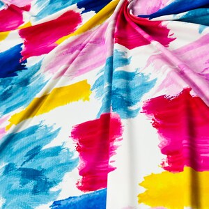 May include: White fabric with a vibrant abstract design. The pattern features broad brushstrokes in shades of blue, pink, red, and yellow. The fabric is folded, creating soft shadows and highlighting the color variations. The overall effect is a modern, artistic textile.