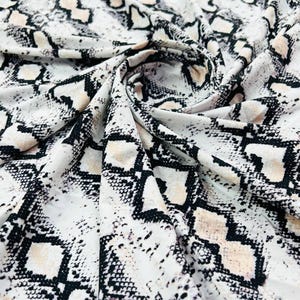 Könnte beinhalten: Weißer Stoff mit einem Schlangenmuster in Schwarz und Beige. Der Stoff ist gefaltet und zeigt die Textur und das Design. Das Muster ist ein sich wiederholendes geometrisches Design.