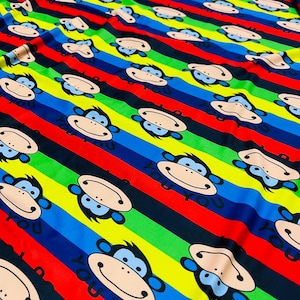 Puede incluir: Tela rayada multicolor con un patrón repetido de monos sonrientes. La tela tiene rayas alternas de rojo, amarillo, verde, azul y negro. Los monos están todos mirando en la misma dirección y tienen ojos azules y un contorno negro.