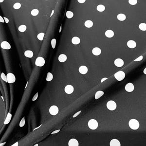 May include: Black fabric with a repeating pattern of large white polka dots. The fabric is draped, showing folds and shadows. The polka dots are evenly spaced and circular, creating a classic and playful design.