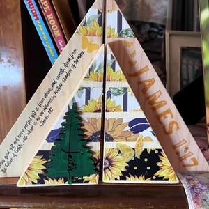 May include: Wooden triangle-shaped decor with sunflower and butterfly designs. One piece has the word "JAMES 1:17" in orange. Another piece has a green tree design and the text "Every good gift...James 1:17".