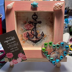 May include: A pink wooden frame with a nautical theme. The frame features an anchor design, seashells, and a heart-shaped embellishment with gems. A card with text from Luke 18:27 is in front of the frame.