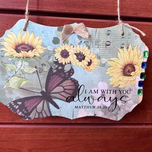 May include: A decorative wooden sign with a floral and butterfly design. The sign features sunflowers, a butterfly, and the text "I AM WITH YOU always" from Matthew 28:20. It has a jute bow and is adorned with colorful gems.
