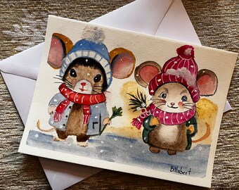 May include: Watercolor greeting card featuring two adorable mice dressed in winter attire. One mouse wears a blue hat and red scarf, the other a red hat and scarf. Both mice are holding sprigs of greenery. The card is on a white envelope.