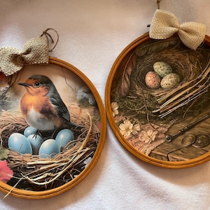 May include: Two round wooden framed decorative pieces. One features a robin sitting on a nest with blue eggs. The other shows a nest with speckled eggs, feathers, and a vintage key. Both have burlap bows.