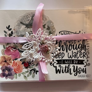 May include: A set of floral-themed greeting cards wrapped in clear cellophane and tied with a light pink ribbon. The cards feature watercolor illustrations of flowers, a bird, and the text "When you pass through deep waters, I will be with you."