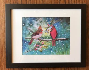 Puede incluir: Obra de arte enmarcada con dos cardenales rojos posados en una rama cubierta de nieve con bayas rojas. El fondo tiene un efecto de acuarela azul y verde con acentos blancos, todo dentro de un marco negro.