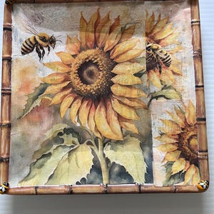 May include: Square decorative tray with a bamboo-style frame. The tray features a watercolor design of sunflowers and bees. The sunflowers are yellow and brown, and the bees are black and yellow. The tray is a decorative item.