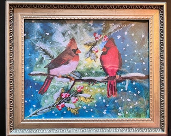 Puede incluir: Una pintura de acuarela enmarcada representa dos cardenales rojos posados en una rama cubierta de nieve con bayas rojas. El fondo presenta una escena invernal nevada, azul y verde. El marco es dorado.