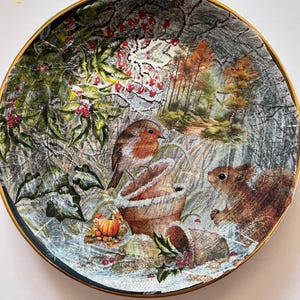 May include: Decorative plate with a winter scene featuring a robin, squirrel, and a pumpkin. The plate has a gold rim and a detailed illustration of a forest with snow and berries. The plate is round.