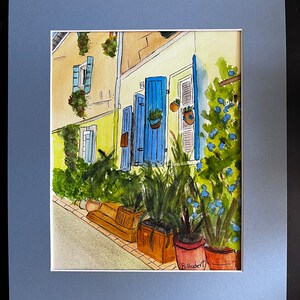 May include: Watercolor painting of a European street scene with yellow and white buildings, blue shutters, and potted plants. The artwork is framed with a blue mat. Signed by B. Hubert.