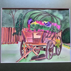 May include: Watercolor painting of a wooden wagon filled with colorful flowers. The wagon has large spoked wheels and is set against a backdrop of a wooden fence, trees, and greenery. The artwork is framed.