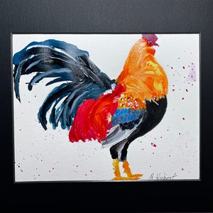 May include: A colorful painting of a rooster, featuring a vibrant orange body, red and blue accents, and a black tail. The artwork is set against a white background with pink paint splatters and is framed in a black mat.