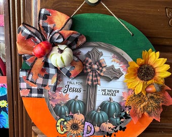 May include: A round, orange and green fall-themed wall hanging with a cross, pumpkins, and a sunflower. The text reads "Fall for Jesus - He Never Leaves" and "All Things Are Possible". A decorative bow and faux pumpkins complete the design.