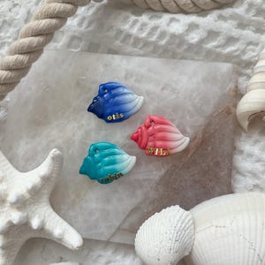 May include: Three seashell-shaped ornaments in blue, pink, and teal with white accents. Each shell has a gold name: "otis", "wila", and "robin". The ornaments are on a marble slab, surrounded by a starfish, rope, and seashells.