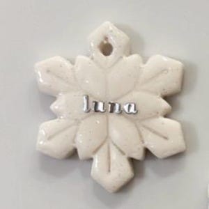 May include: A white snowflake-shaped ornament with the name "Luna" in silver letters. The ornament has a glittery finish and a hole at the top for hanging. The snowflake design features six points with leaf-like details.