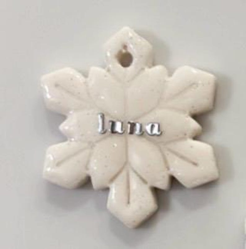 May include: A white snowflake-shaped ornament with the name "Luna" in silver letters. The ornament has a glittery finish and a hole at the top for hanging. The snowflake design features six points with leaf-like details.