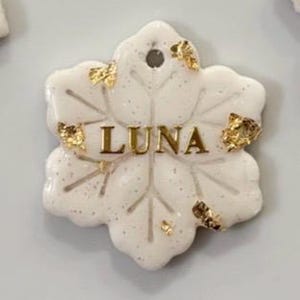 May include: A white, snowflake-shaped tag with the name "LUNA" in gold lettering. The tag is speckled with glitter and adorned with gold leaf accents. It has a small hole at the top for attachment.