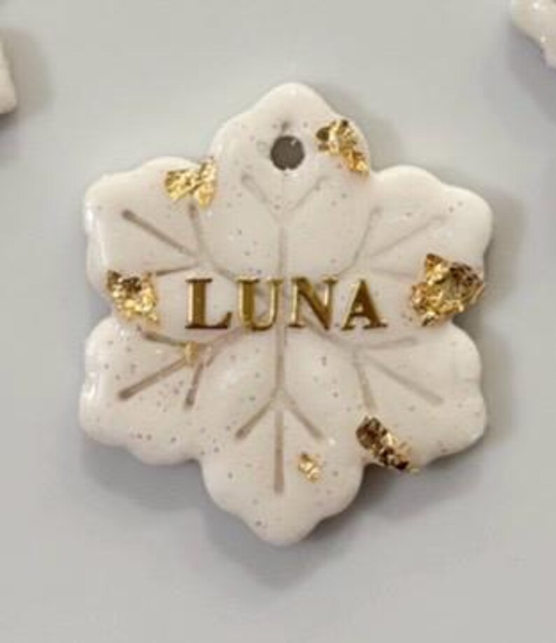 May include: A white, snowflake-shaped tag with the name "LUNA" in gold lettering. The tag is speckled with glitter and adorned with gold leaf accents. It has a small hole at the top for attachment.