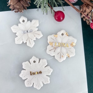 May include: Three white snowflake ornaments with the name "Luna" in silver and gold lettering. The ornaments have a glittery finish and are displayed on a white marble surface. Decorative holiday theme.