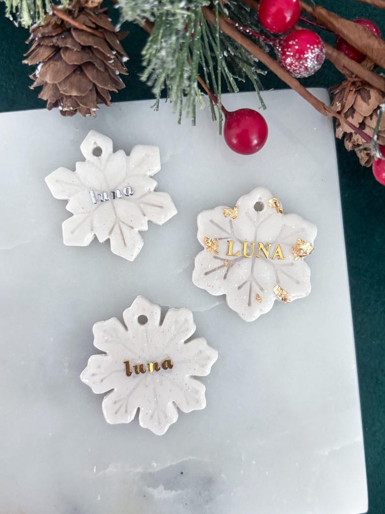 May include: Three white snowflake ornaments with the name "Luna" in silver and gold lettering. The ornaments have a glittery finish and are displayed on a white marble surface. Decorative holiday theme.