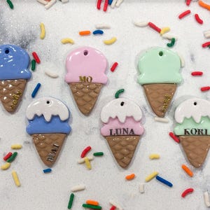 以下が含まれることがあります： 「MO」や「LUNA」などの名前が書かれた、パステルカラーのアイスクリームコーンのチャームのコレクション。チャームは青、ピンク、緑で、コーンは茶色。カラフルなスプリンクルがチャームを囲んでいます。