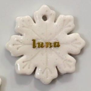 May include: A white snowflake-shaped ornament with the name "Luna" in gold lettering. The ornament has a glossy finish and a small hole at the top for hanging. The snowflake design features six points with detailed edges.