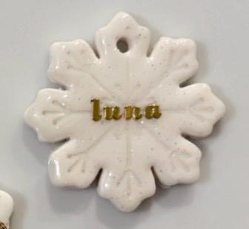 May include: A white snowflake-shaped ornament with the name "Luna" in gold lettering. The ornament has a glossy finish and a small hole at the top for hanging. The snowflake design features six points with detailed edges.