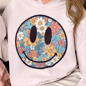 May include: A white hoodie with a floral smiley face design. The hoodie has a drawstring hood and long sleeves.