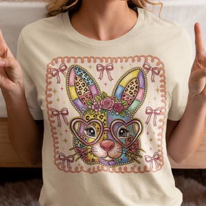 Può includere: T-shirt color crema con un design a forma di coniglio colorato. Il coniglio ha occhiali a forma di cuore, accenti floreali e orecchie patchwork. Il design è incorniciato da fiocchi rosa e un bordo smerlato, perfetto per Pasqua.