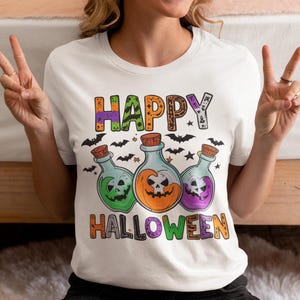 May include: White t-shirt with a Halloween-themed graphic. The design features three potion bottles with smiling skull faces in green, orange, and purple, with the words "HAPPY HALLOWEEN" above and below. Bats and stars are also included.