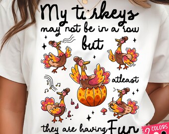 Pode incluir: T-shirt branco com um gráfico colorido de desenhos animados de perus e o texto "My turkeys may not be in a row but at least they are having fun." O design inclui notas musicais e estrelas.
