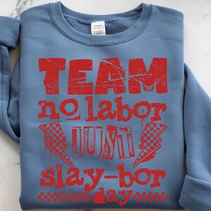 Może przedstawiać: Niebieska bluza z napisem "TEAM no labor JUST slay-bor day" w postarzanym czerwonym druku. Projekt zawiera szachownicowe błyskawice. Bluza jest złożona na białej powierzchni.