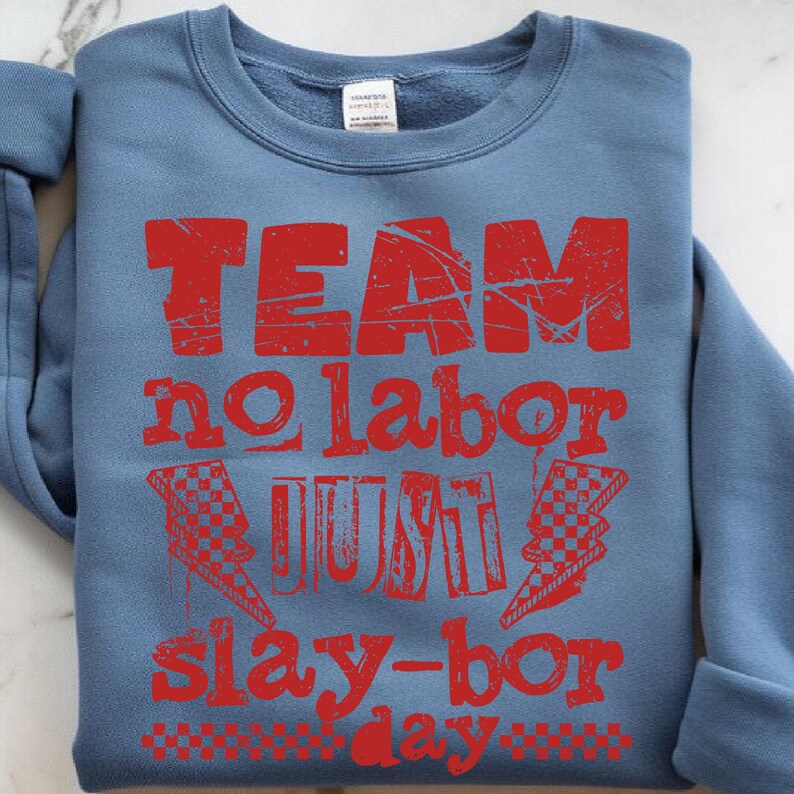 Może przedstawiać: Niebieska bluza z napisem "TEAM no labor JUST slay-bor day" w postarzanym czerwonym druku. Projekt zawiera szachownicowe błyskawice. Bluza jest złożona na białej powierzchni.